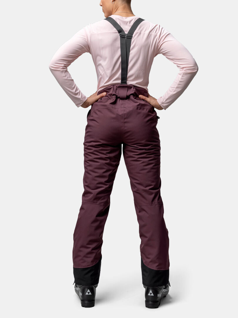 Eine Person in Halti Carvey II DX Skihose Damen mit schwarzen Hosenträgern und Skischuhen steht bereit zum Skifahren.