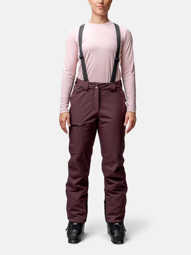 Eine Person steht vorne in einem rosa Hemd, Halti Carvey II DX Skihose Damen mit Hosenträgern und schwarzen Stiefeln.