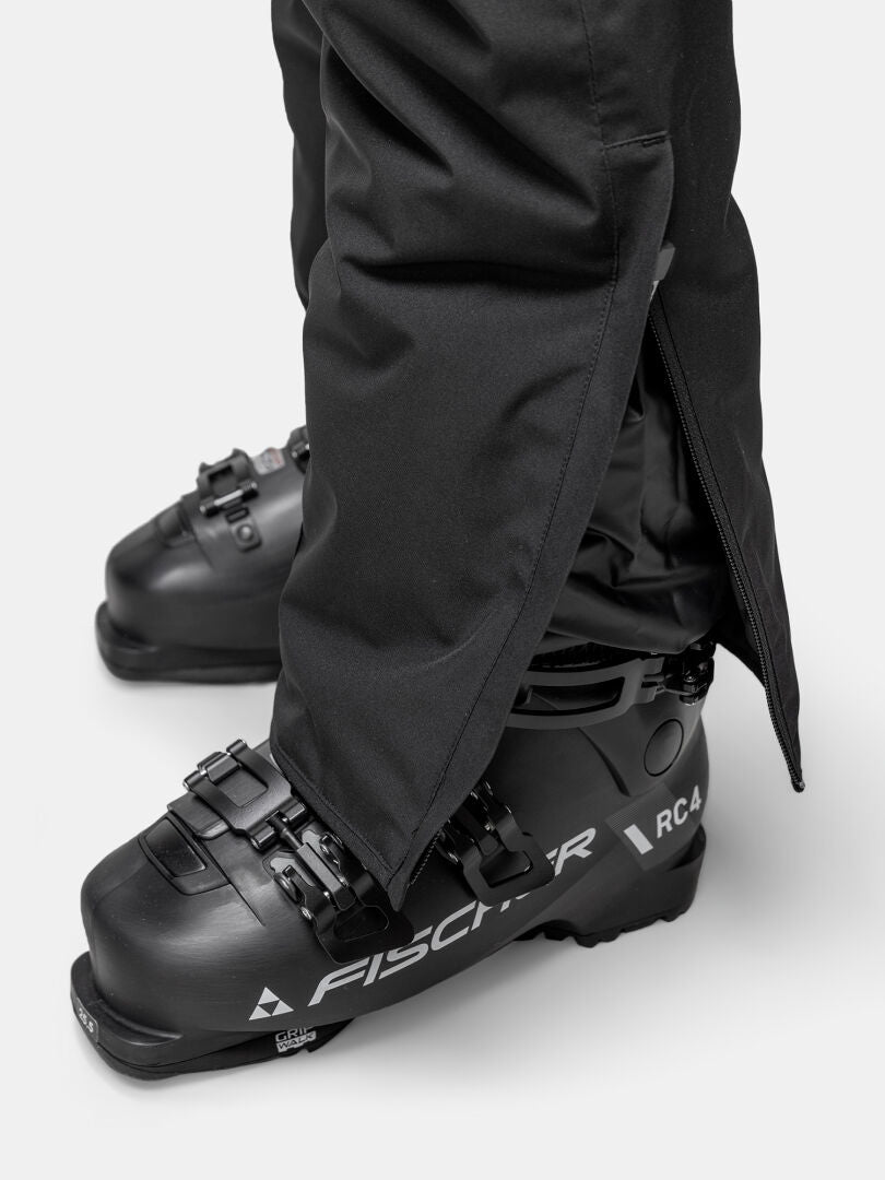 Eine Person in Halti Glades II DX Skihose Damen und schwarzen Fischer RC4 Skischuhen, die Hose ist an den Knöcheln offen.