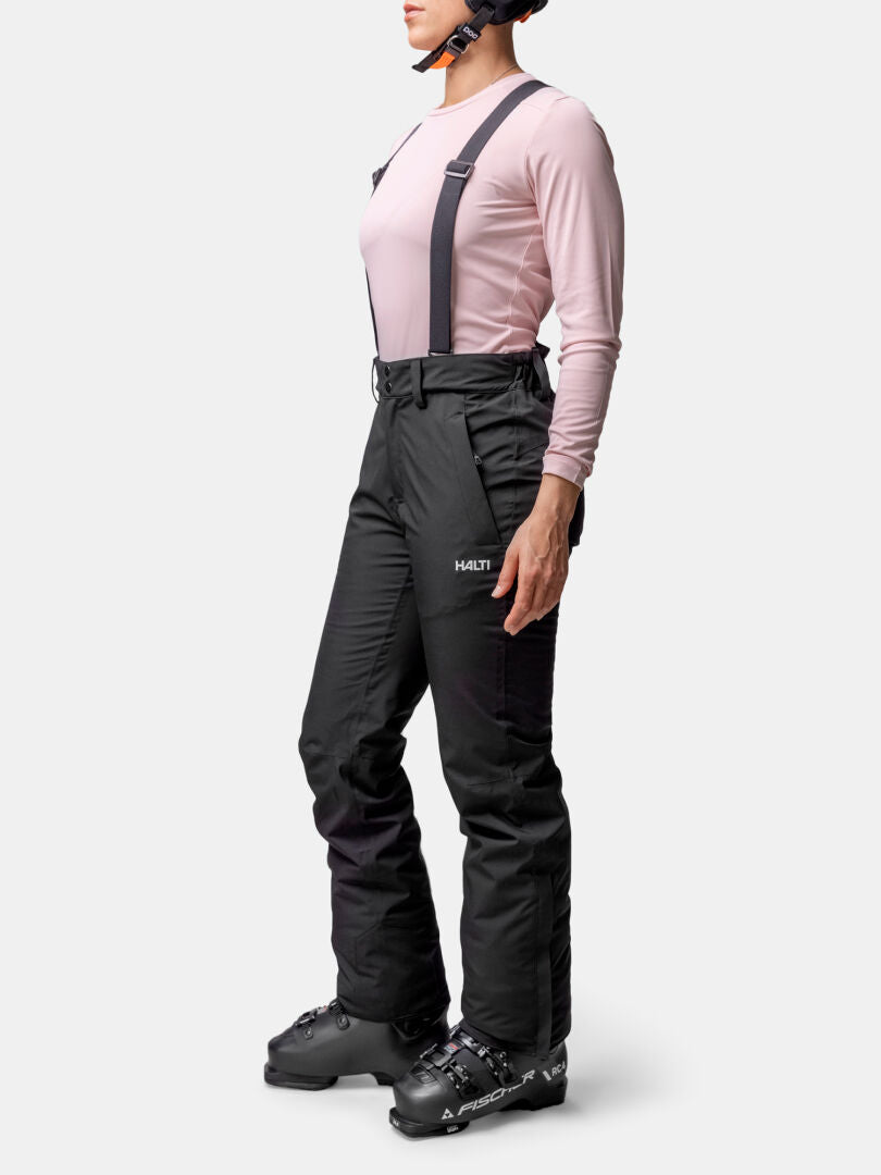 Eine Person in Halti Glades II DX Skihose Damen, rosa Langarmshirt, Skischuhen und Helm steht seitlich auf der Piste.