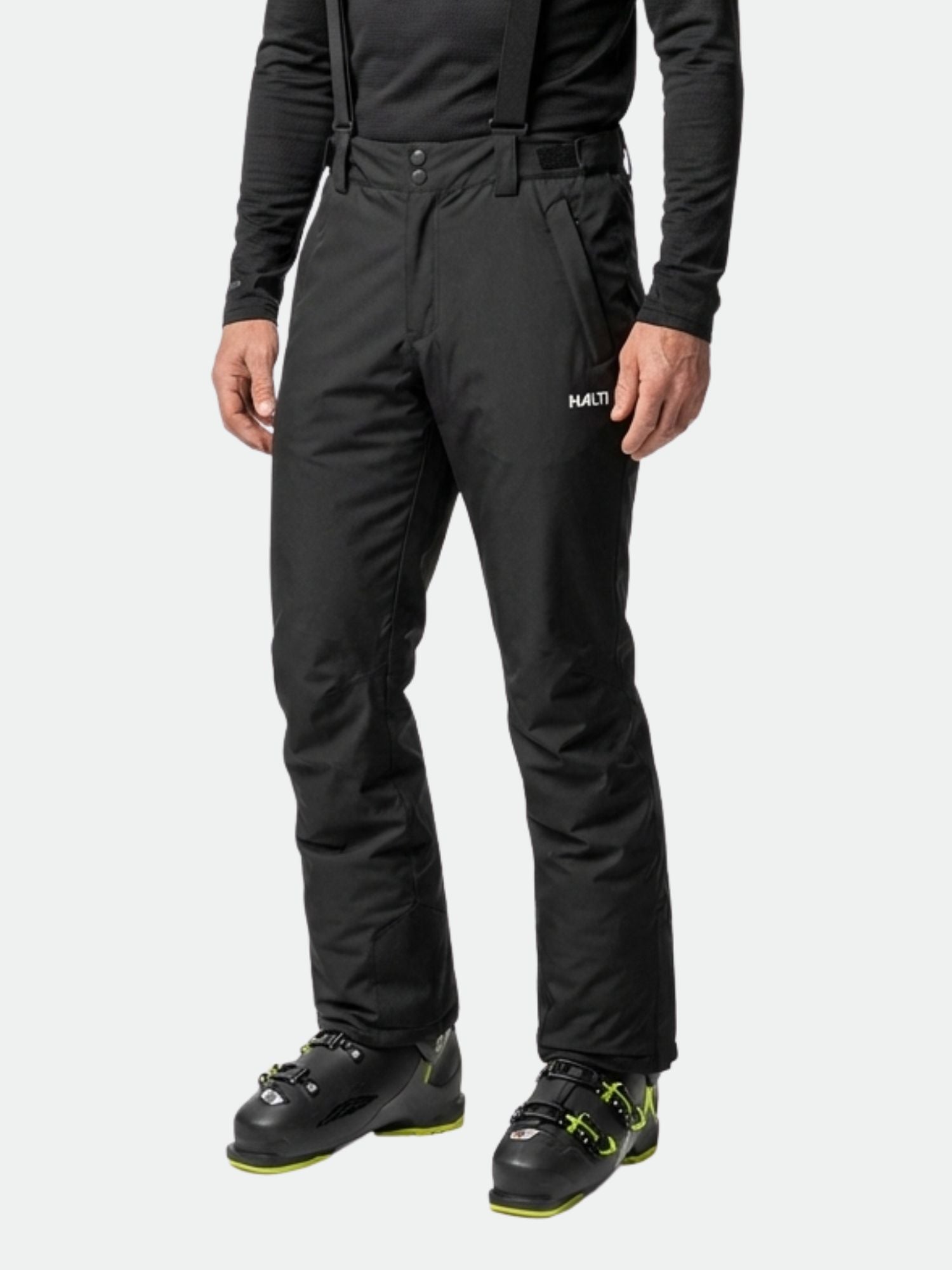 Eine Person in Halti Glades II DX Skihose Herren, schwarzen Stiefeln mit grünen Akzenten und einem schwarzen langärmeligen Hemd.