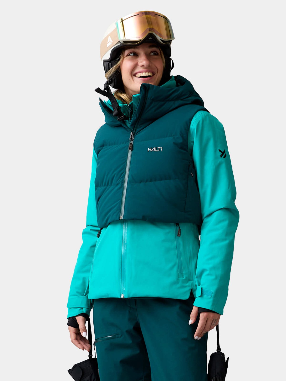 Eine lächelnde Frau trägt eine Halti Varion 2in1 DX Skijacke für Damen, einen Helm und eine Skibrille sowie eine abnehmbare dunkelblaugrüne Weste.