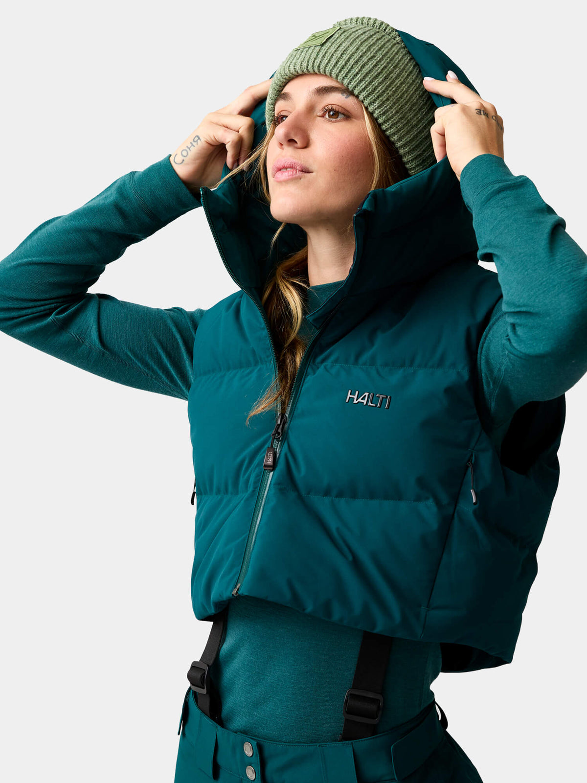 Eine Frau in einer dunkelblaugrünen Halti Varion 2in1 DX Skijacke für Damen und einer grünen Strickmütze.