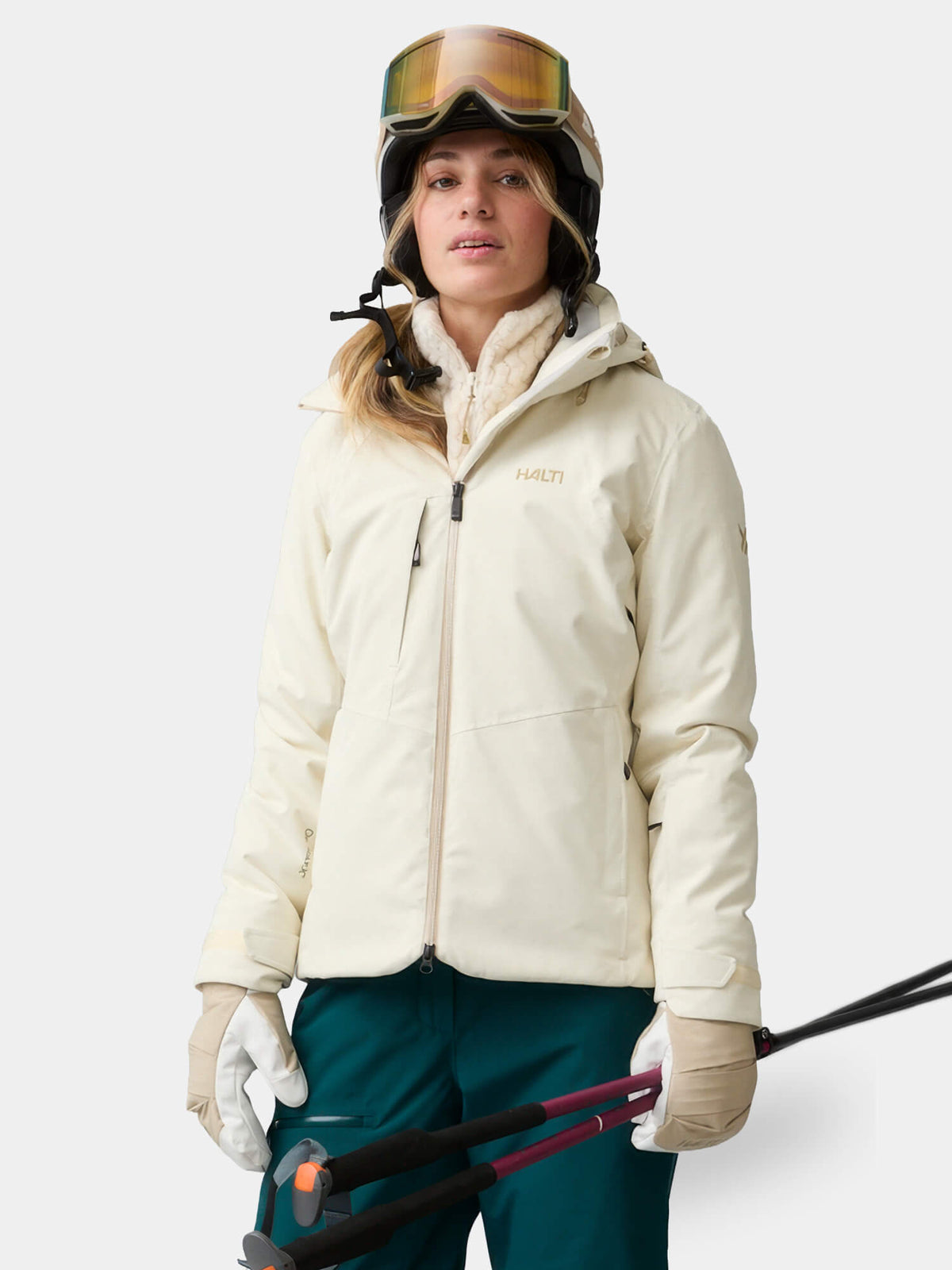 Eine Frau in einer cremefarbenen Halti Recier DX Skijacke, die einen Helm und eine Skibrille trägt und Skistöcke in den Händen hält.