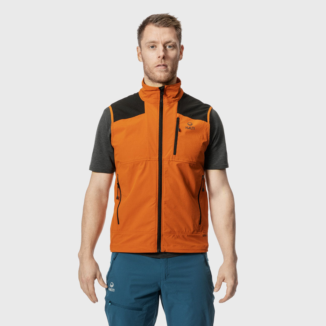 Ein Mann, der eine leuchtend orangefarbene Weste mit Reißverschluss über einem dunklen T-Shirt und einer blauen Hose trägt, steht mit dem Gesicht nach vorne vor einem einfarbigen Hintergrund.