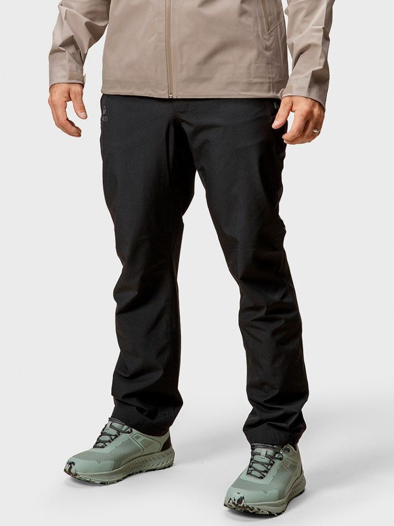 Eine Person in beiger Jacke, Halti Vuoksi DX Regenhose Herren und hellgrünen Wanderschuhen steht vor einem schlichten Hintergrund.