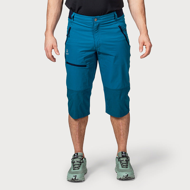Eine Person, die knielange, blaugrüne Outdoor-Shorts, ein schwarzes Hemd und grüne Wanderschuhe trägt, steht vor einem weißen Hintergrund.