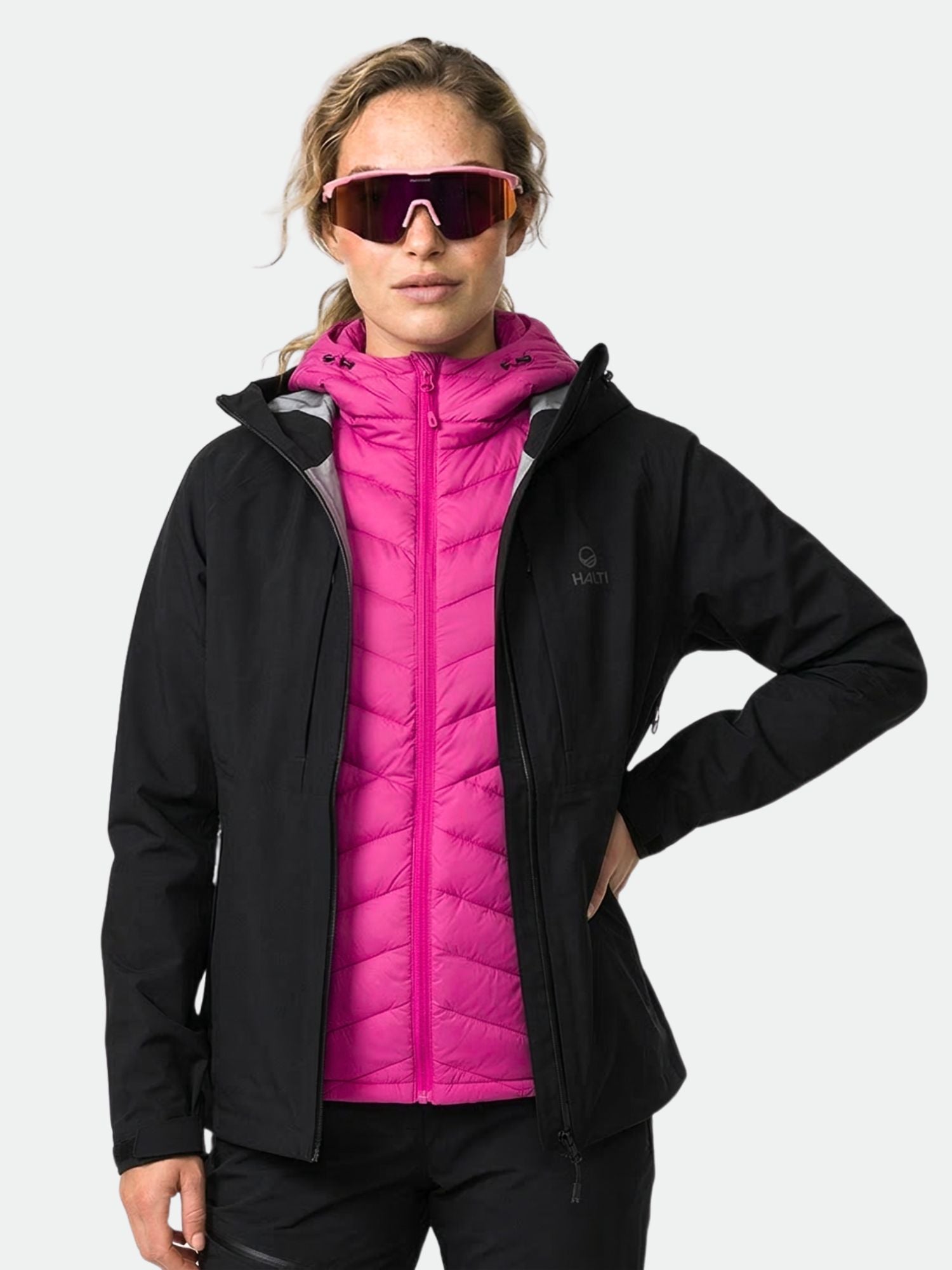 Eine Person in einer Halti Kaarna Damen DX 3L Regenjacke über einer rosafarbenen Puffjacke und einer reflektierenden Sonnenbrille steht vor einem unifarbenen Hintergrund.