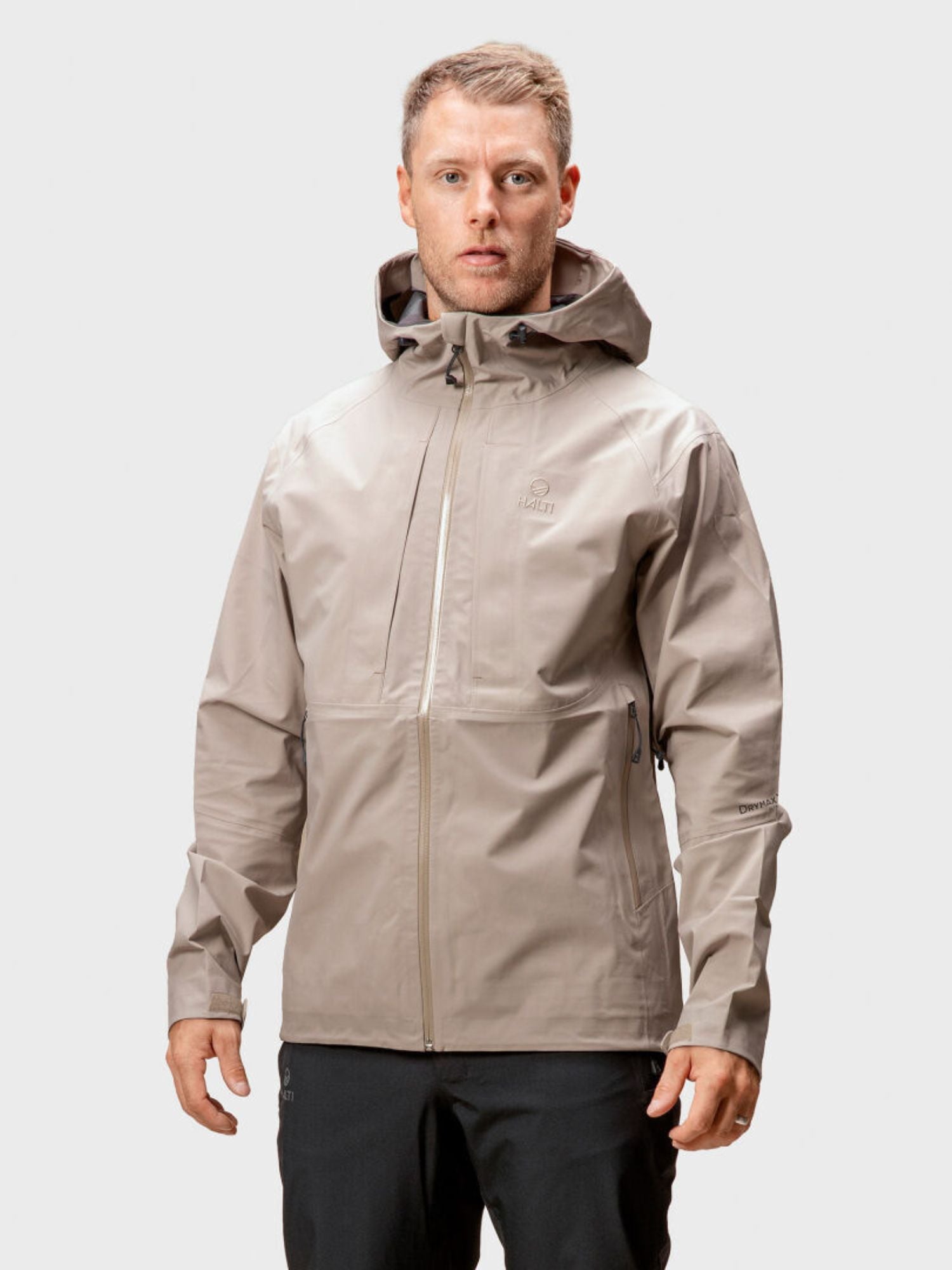 Ein Mann in einer hellgrauen Halti Kaarna Herren DrymaxX 3L Regenjacke und schwarzer Hose steht vor einem schlichten Hintergrund.