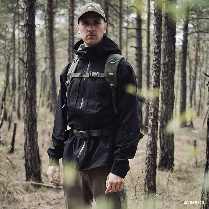 Ein Mann in einer Halti Kaarna Herren DrymaxX 3L Regenjacke mit Rucksack steht in einem Wald, bereit für Outdoor-Aktivitäten.