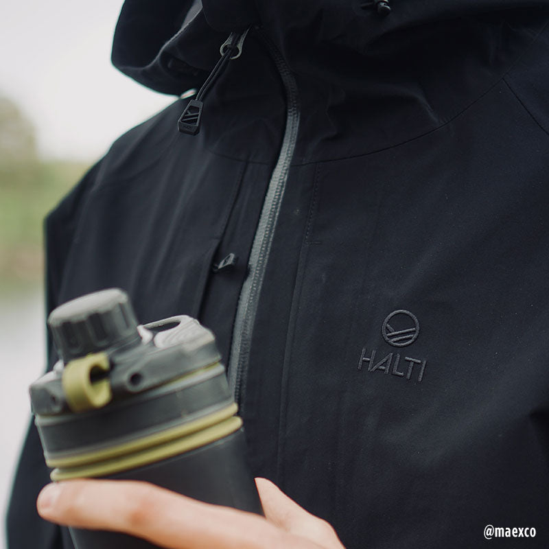 Person trägt die Halti Kaarna Herren DrymaxX 3L Regenjacke und hält draußen eine schwarz-gelbe Wasserflasche.