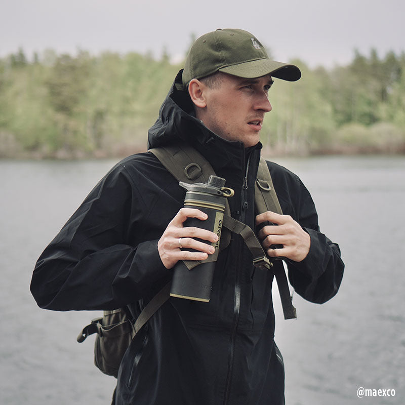 Ein Mann in Outdoor-Ausrüstung trägt eine Halti Kaarna Herren DrymaxX 3L Regenjacke und hält einen Becher an einem See mit Bäumen dahinter.