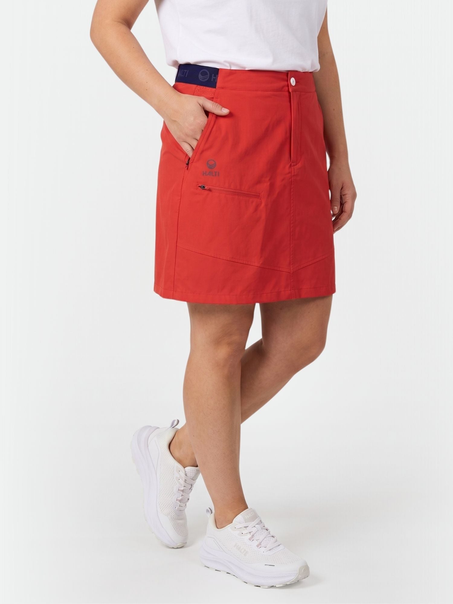 Eine Person trägt den Halti Pallas Damen Lite X-stretch Rock in rot, ein weißes T-Shirt und Turnschuhe, die Hand in der Skorttasche.