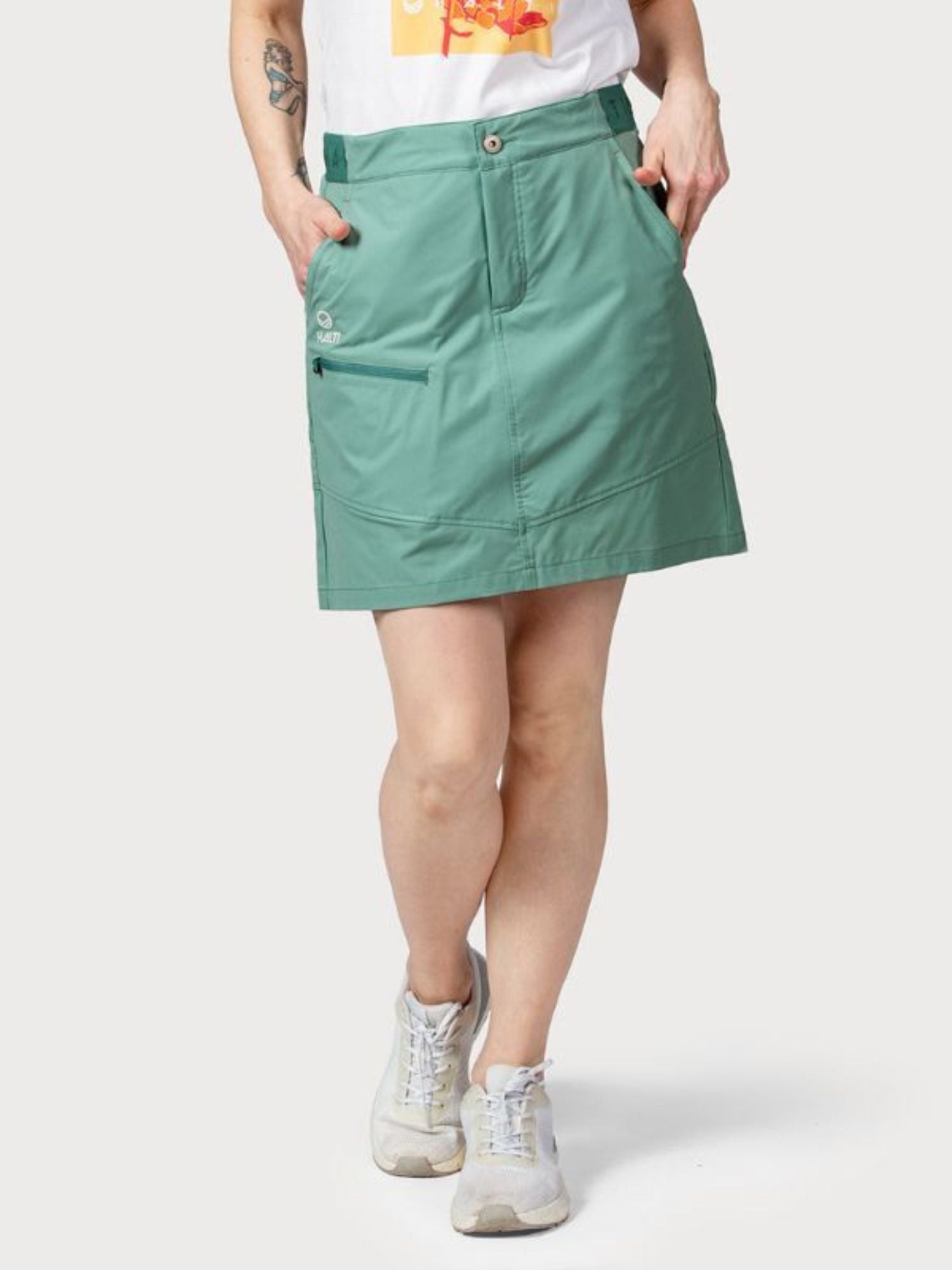 Person trägt einen grünen Halti Pallas Damen Lite X-Stretch Rock, ein weißes T-Shirt mit Grafik und Turnschuhe, Hände in den Taschen.