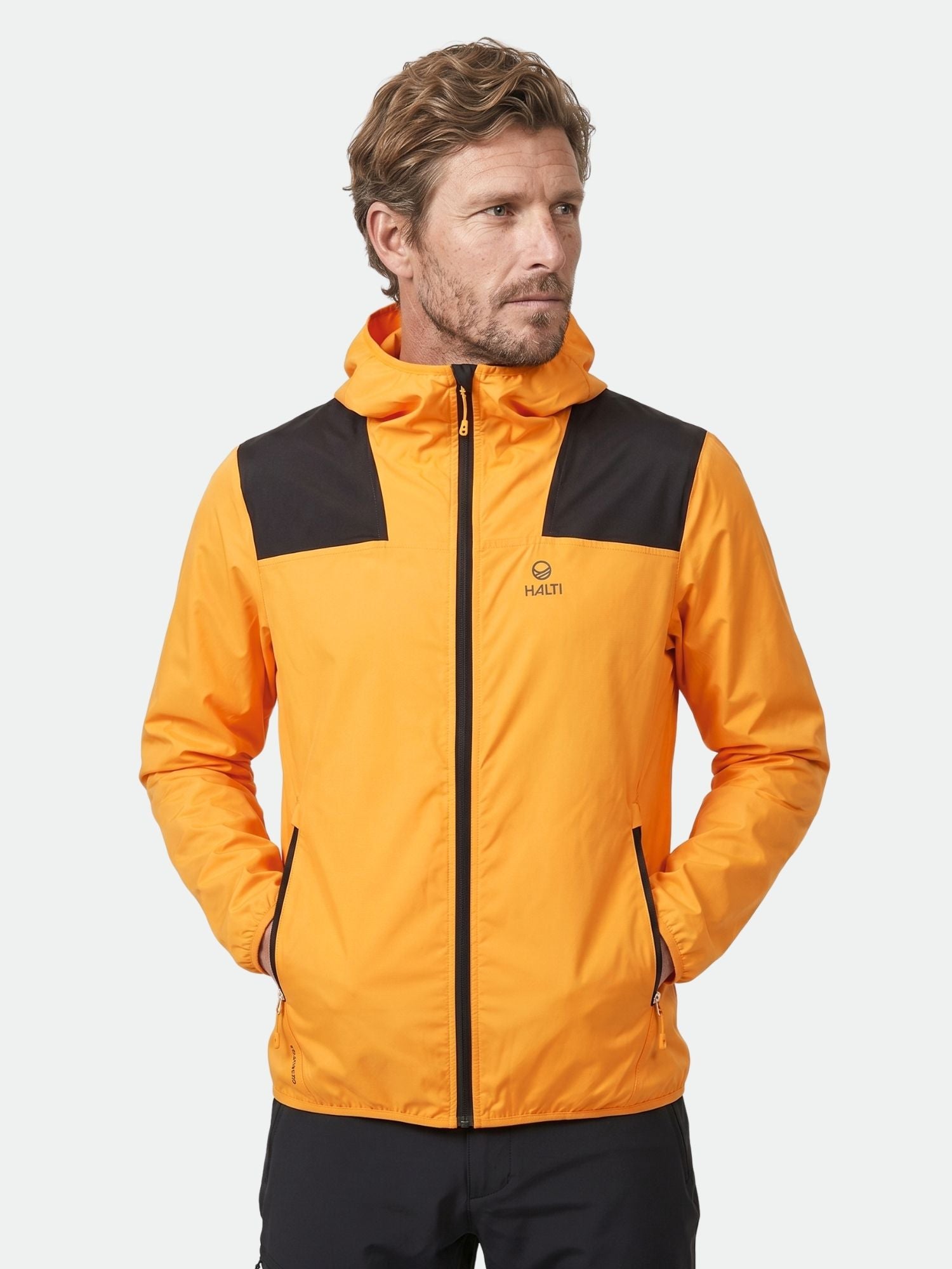 Mann in Halti Pallas Herren Lite X-stretch Jacke, orange/schwarz, Hände in den Taschen, vor einem schlichten hellen Hintergrund.