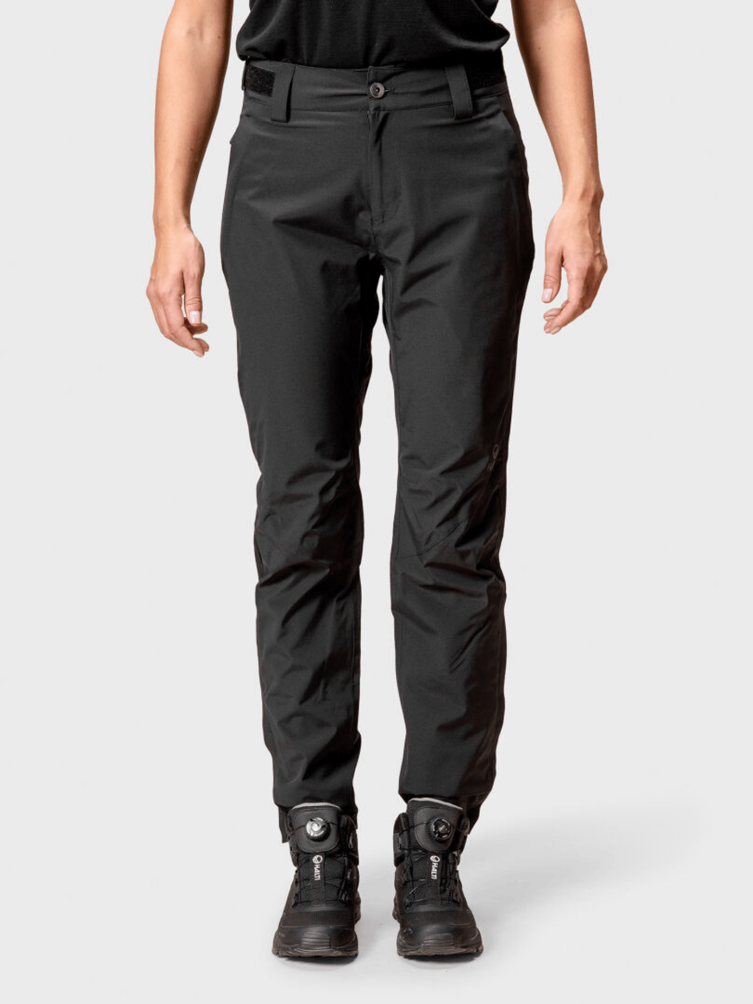 Eine Person trägt Halti Juonto Damen Regenhose und Wanderstiefel und steht vor einem schlichten hellen Hintergrund.