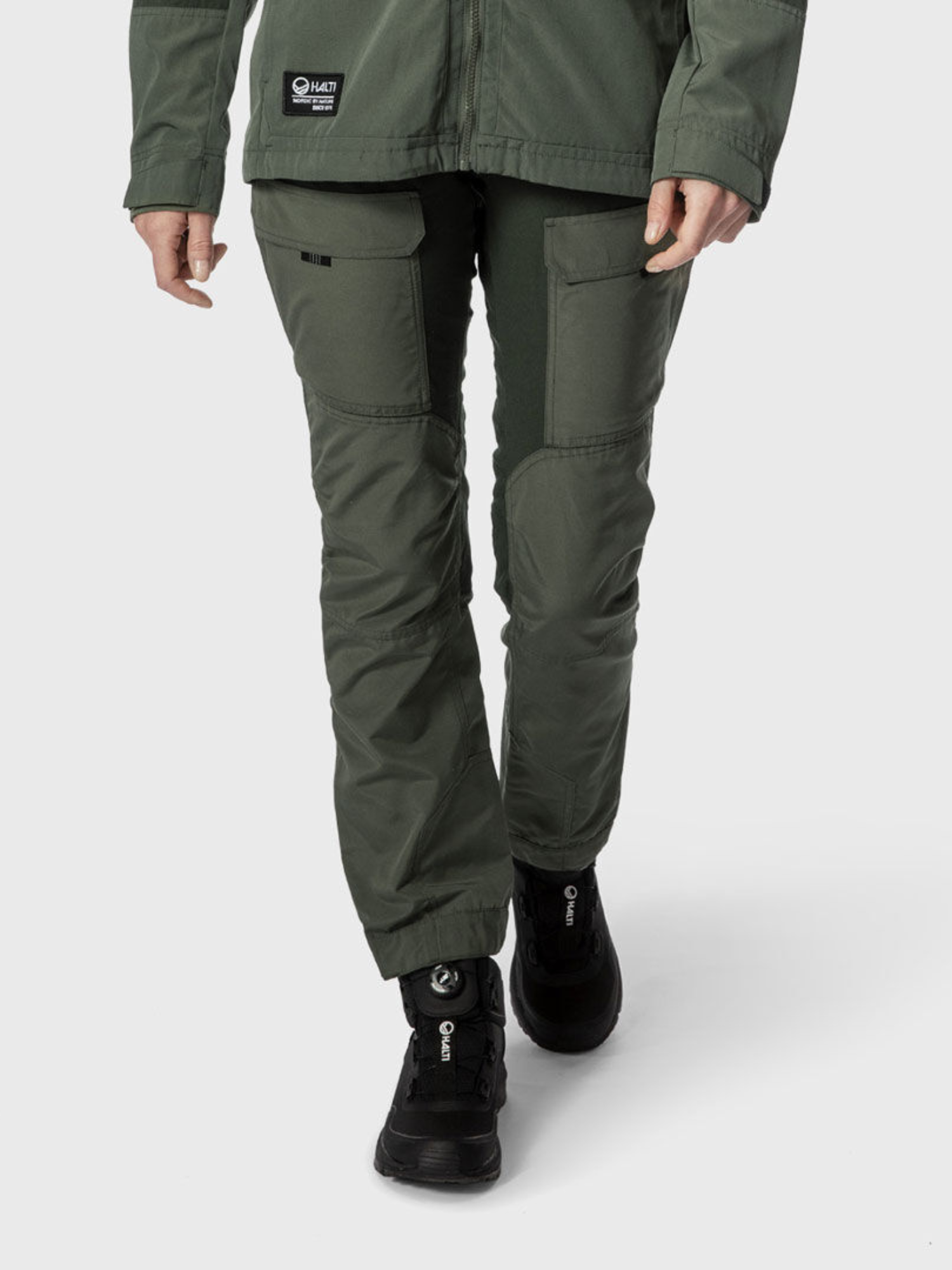 Eine Person trägt Halti Hiker Damen Ventilated Hose in grünen und schwarzen Wanderschuhen, dargestellt von der Taille bis zu den Füßen auf einfarbigem Hintergrund.