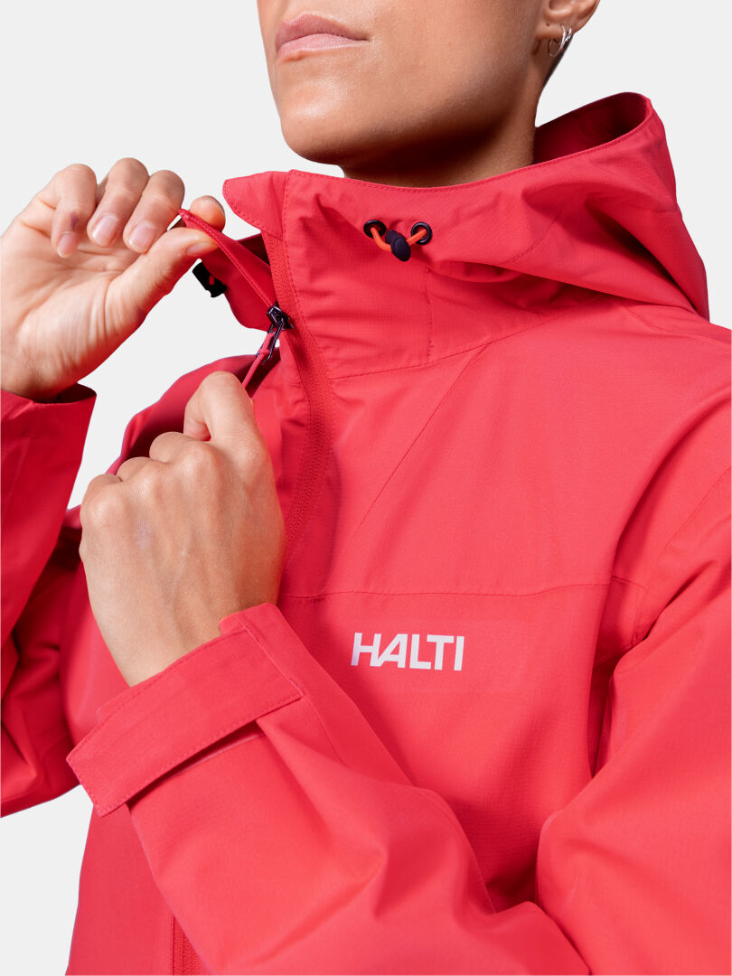 Eine Person schließt den Reißverschluss einer roten Halti Forter DX Regenjacke Damen, die das Logo zeigt - ideal für Aktivitäten im Freien.