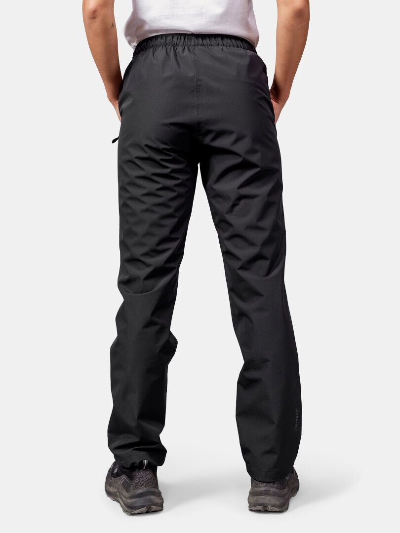 Eine Person in Halti Forter DX Regenhose Damen und dunklen Schuhen steht mit dem Gesicht nach unten, die Hände in den Taschen, vor einem hellen Hintergrund.