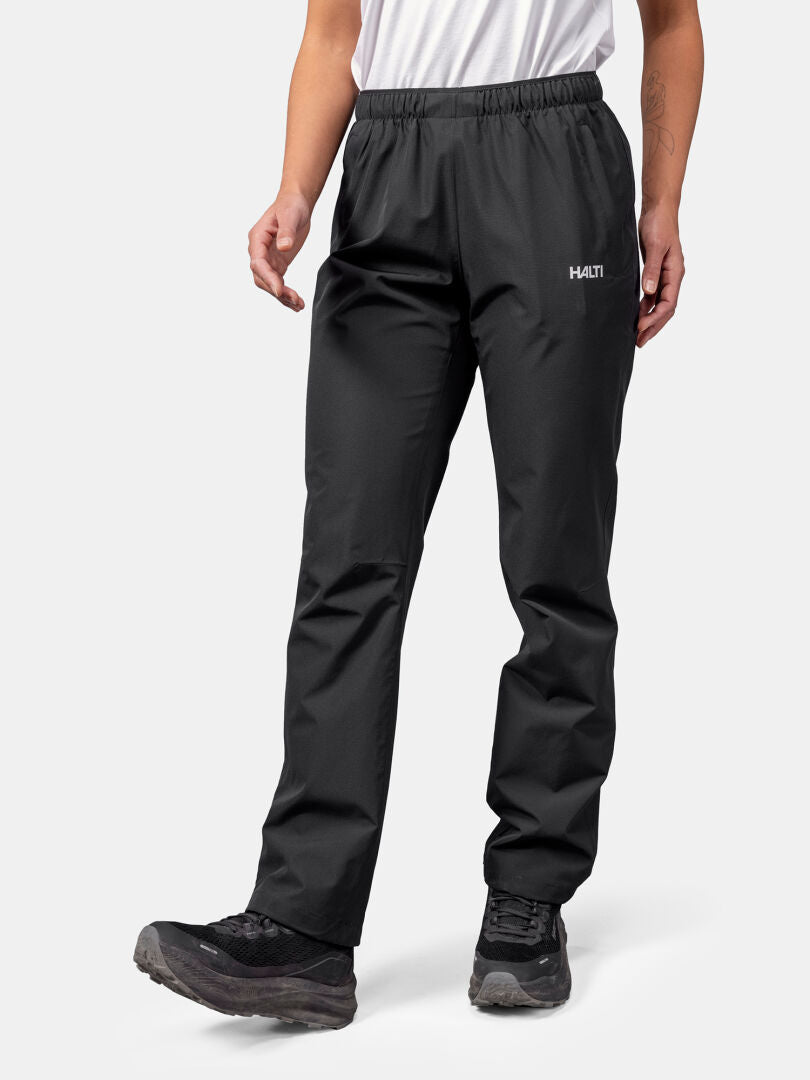 Eine Person in schwarzen Halti Forter DX Regenhose Damen und Wanderschuhen, die ein weißes Hemd trägt, steht vor einem schlichten Hintergrund.