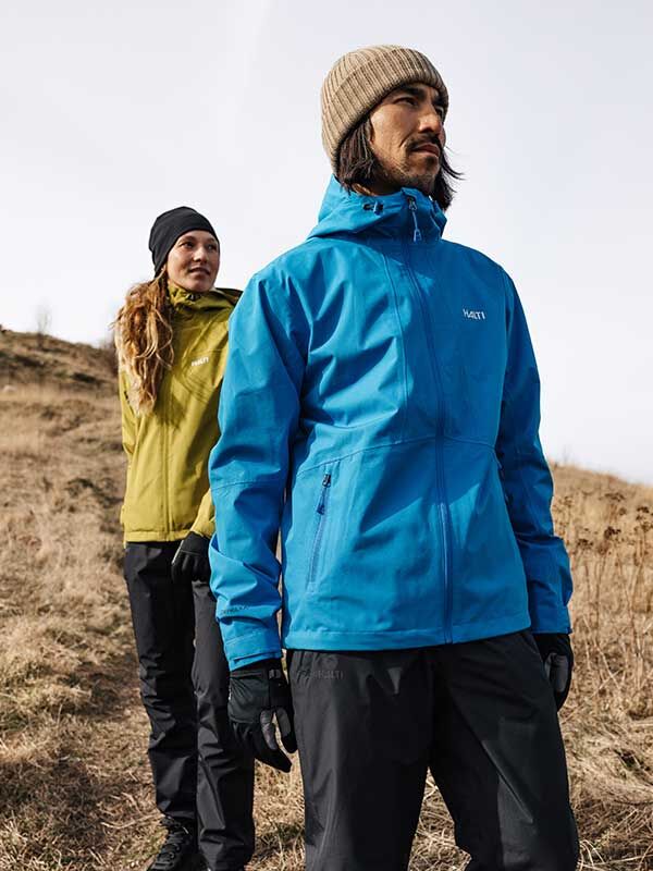Zwei Personen tragen die Halti Forter DX Herren-Regenjacke und stehen an einem bewölkten Tag beim Wandern auf einer grünen Anhöhe.