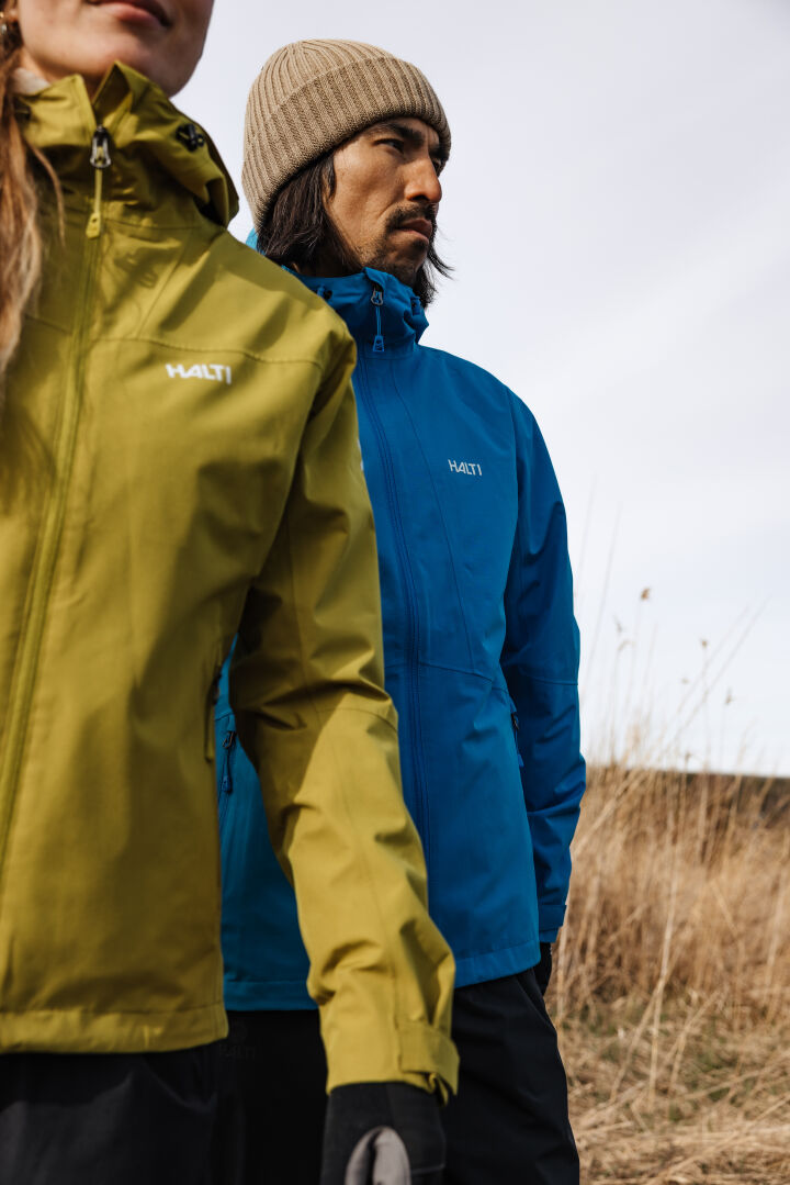 Zwei Personen in der Halti Forter DX Regenjacke für Herren – eine in Gelbgrün und eine in Blau – stehen auf einer Wiese, bereit für eine Wanderung.