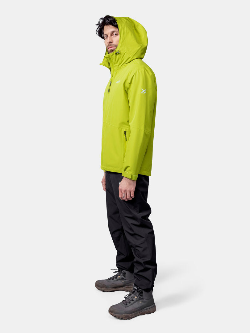 Ein Mann steht seitlich in einer leuchtend grünen Halti Forter DX Regenjacke Herren, einer schwarzen Hose und grauen Wanderschuhen.