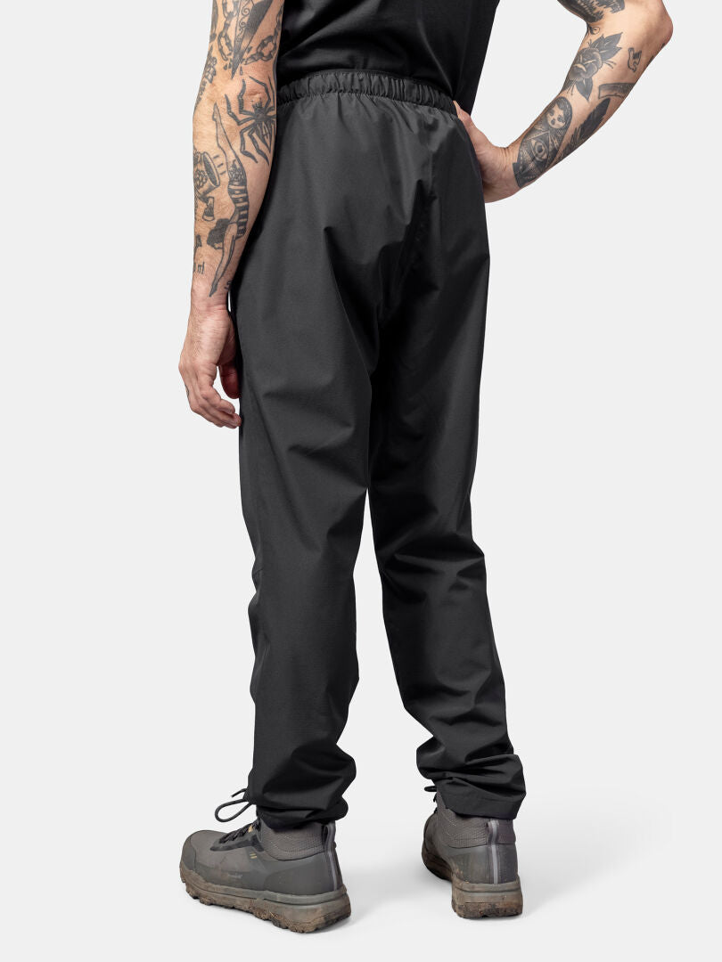 Eine Person in Halti Forter DrymaxX Regenhose Herren schwarzer Hose, grauen Stiefeln und schwarzem Hemd steht mit dem Rücken zur Wand.