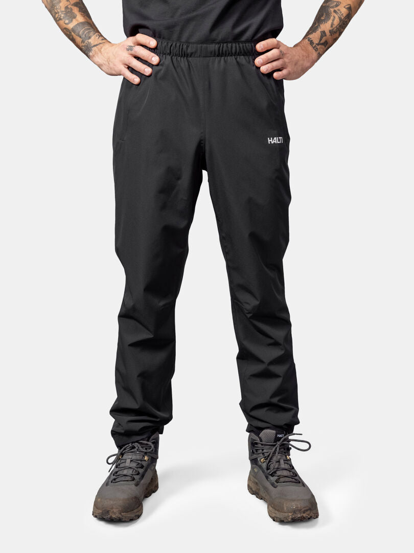 Mann mit Halti Forter DrymaxX Regenhose, schwarzer Hose und Wanderschuhen, zeigt Armtattoos auf weißem Hintergrund.