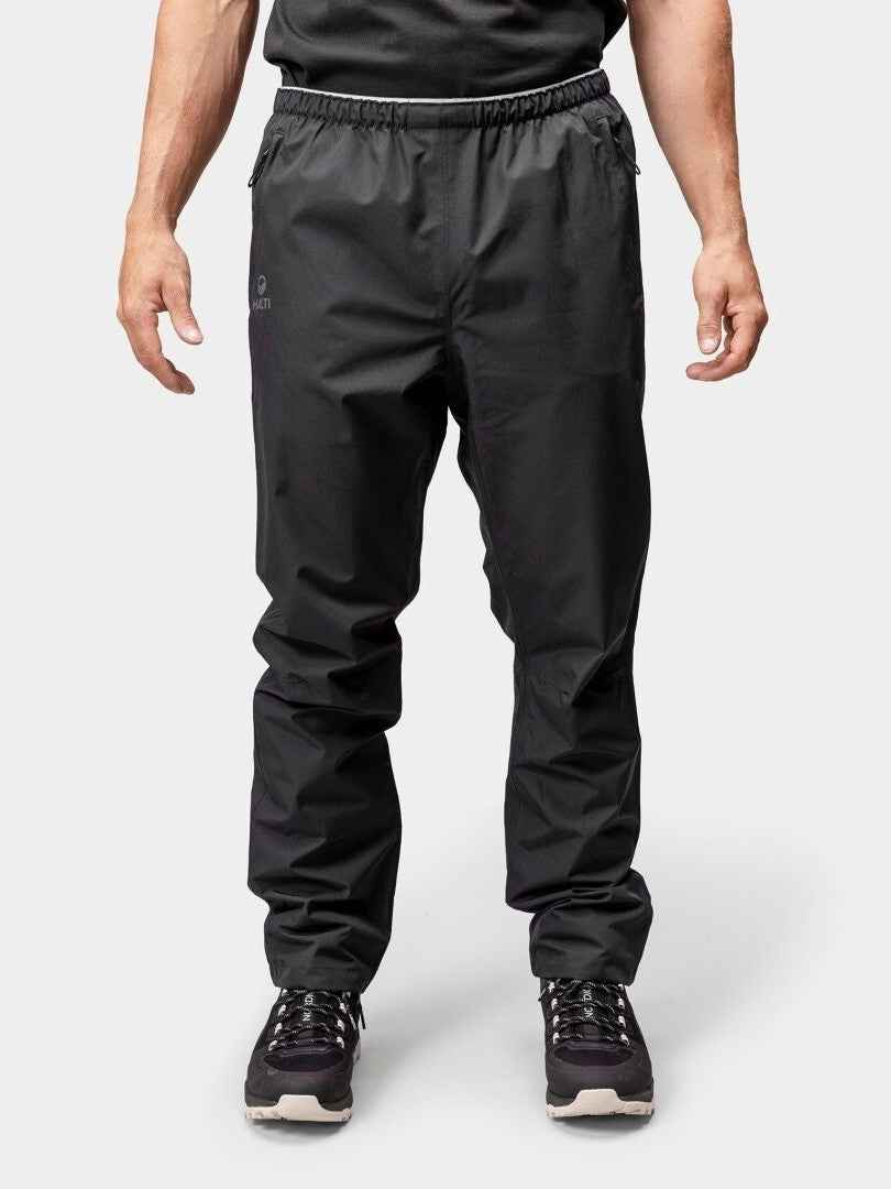 Eine Person in Halti Forter DX Regenhose Herren und Turnschuhen steht vor einem hellen Hintergrund, bereit zum Wandern oder Regen.