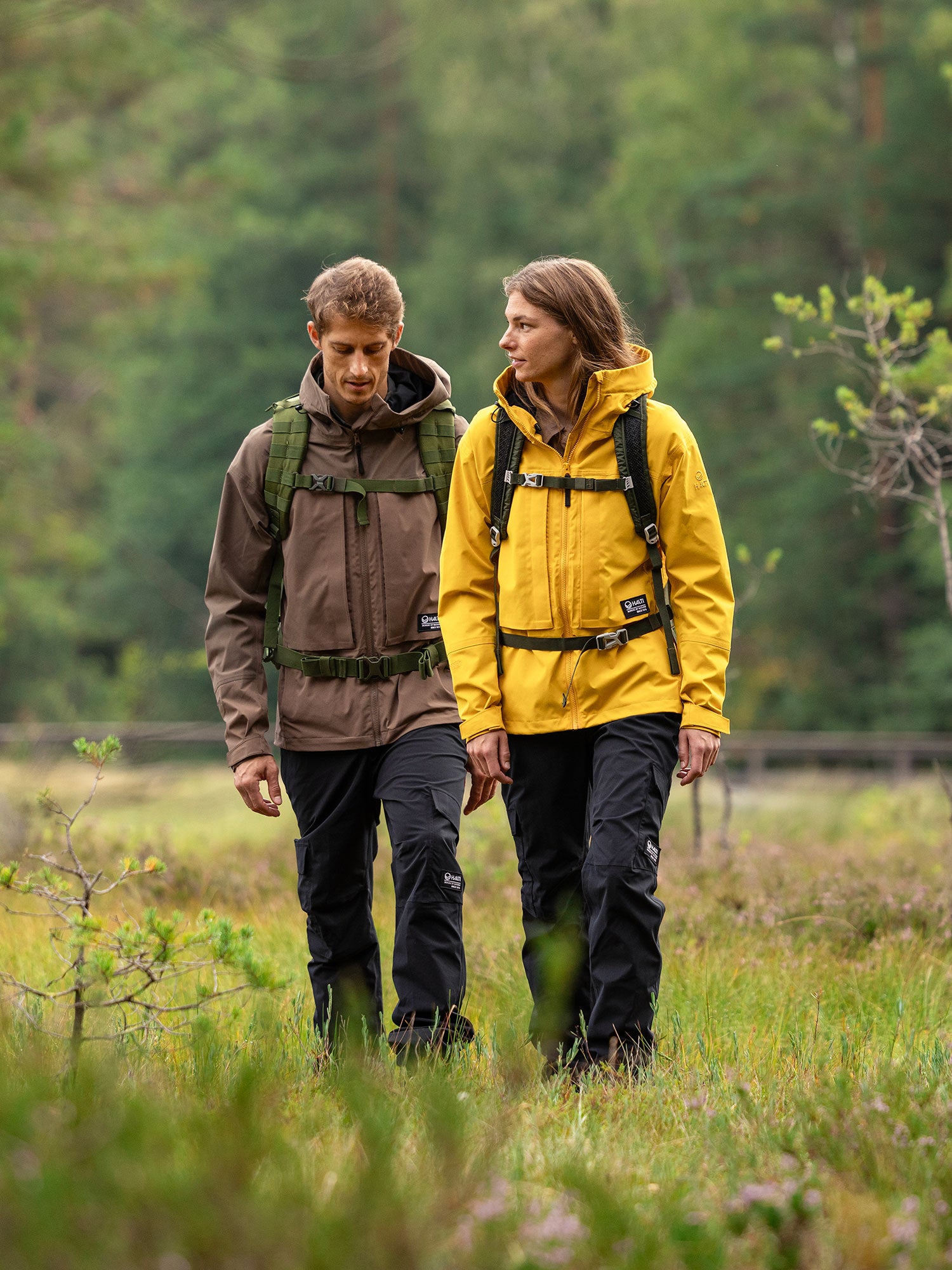 Zwei Personen in Halti Hiker II DX Regenjacke Damen und Rucksäcken gehen gemeinsam auf einem grasbewachsenen Weg mit Bäumen im Hintergrund.