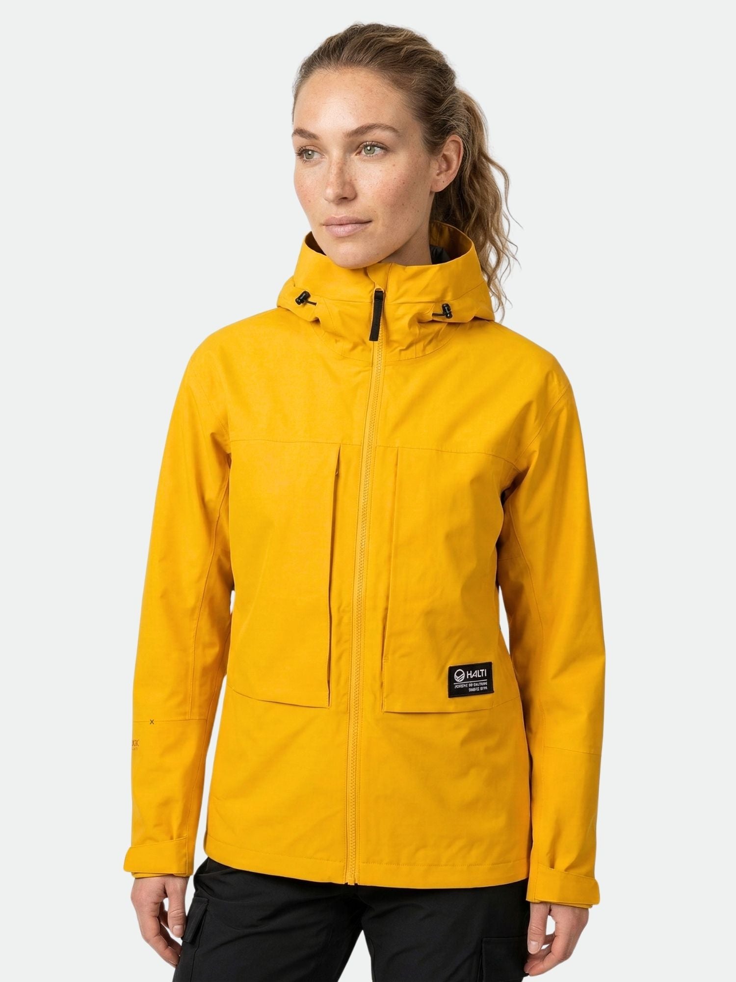 Eine Person, die die leuchtend gelbe Halti Hiker II DX Regenjacke Damen trägt, steht vor einem schlichten hellen Hintergrund.