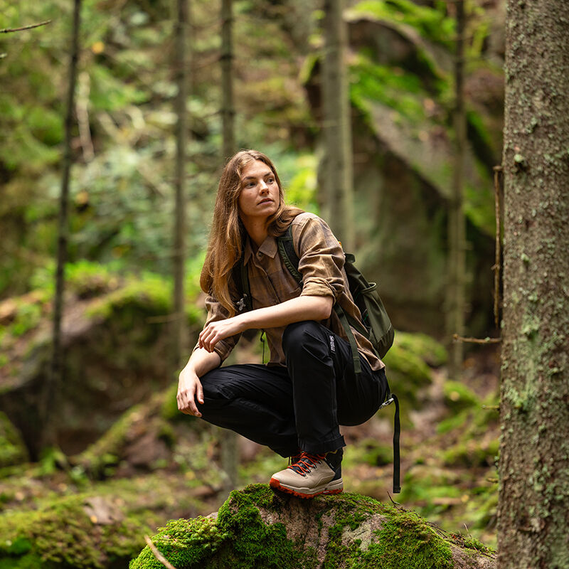Eine Person mit langen Haaren hockt auf einem moosigen Felsen in einem grünen Wald und trägt Halti Hiker II DrymaxX Hosen Damen und einen Rucksack.