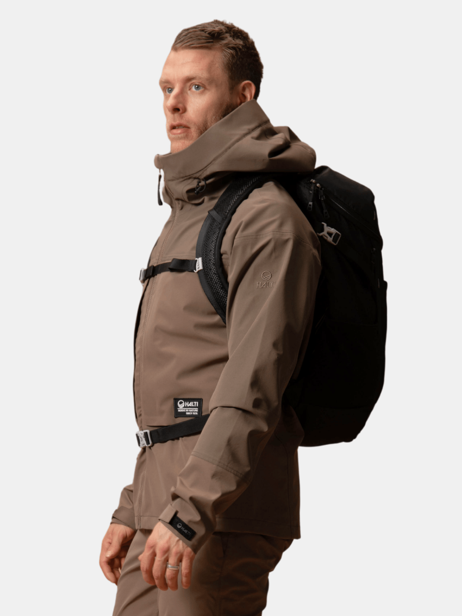 Eine Person in einer Halti Hiker II DrymaxX Regenjacke für Herren steht seitlich mit einem schwarzen Rucksack, einfarbiger Hintergrund.