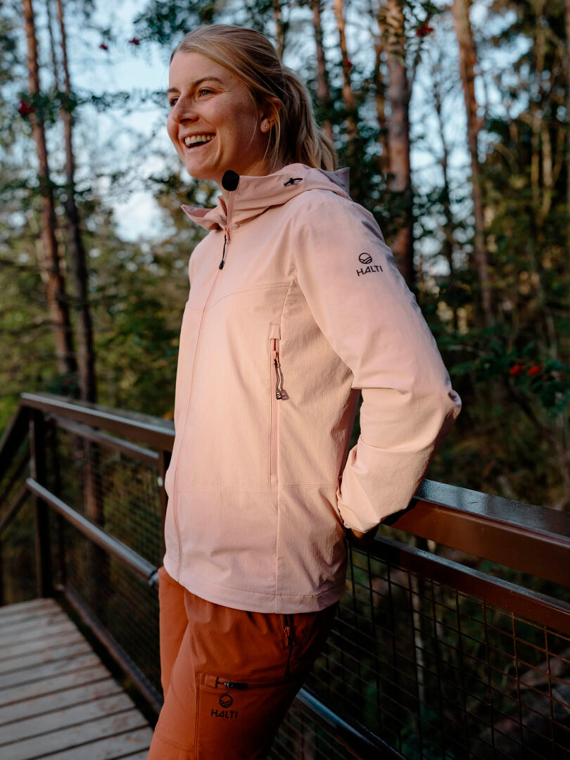 Eine Frau in einer Halti Pallas Evo X-Stretch-Jacke und orangefarbenen Hosen lächelt auf einer Holzbrücke im Wald.