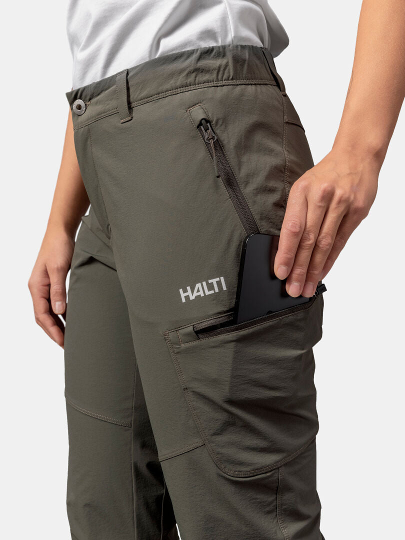 Eine Person steckt ein schwarzes Smartphone in die Oberschenkeltasche mit Reißverschluss der olivgrünen Halti Pallas Evo X-Stretch-Hosen Damen.