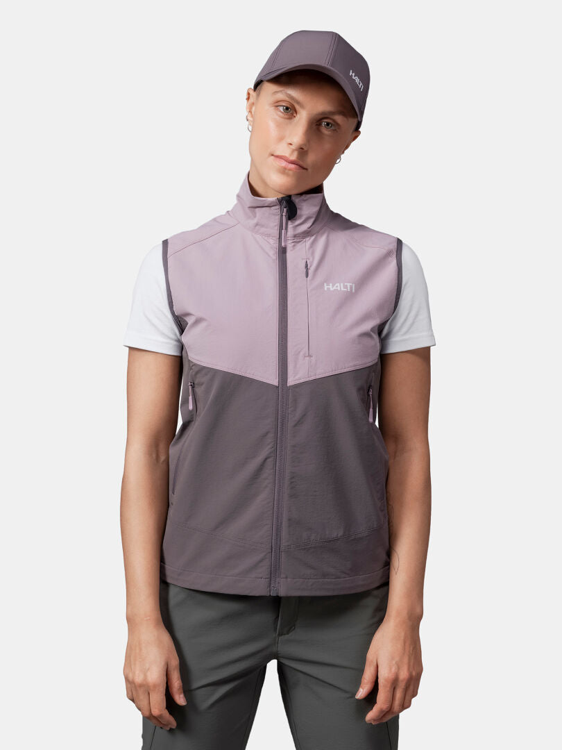Eine Person trägt die Halti Pallas Evo X-stretch Weste Damen, ein weißes T-Shirt und eine Mütze vor einem einfarbigen Hintergrund.