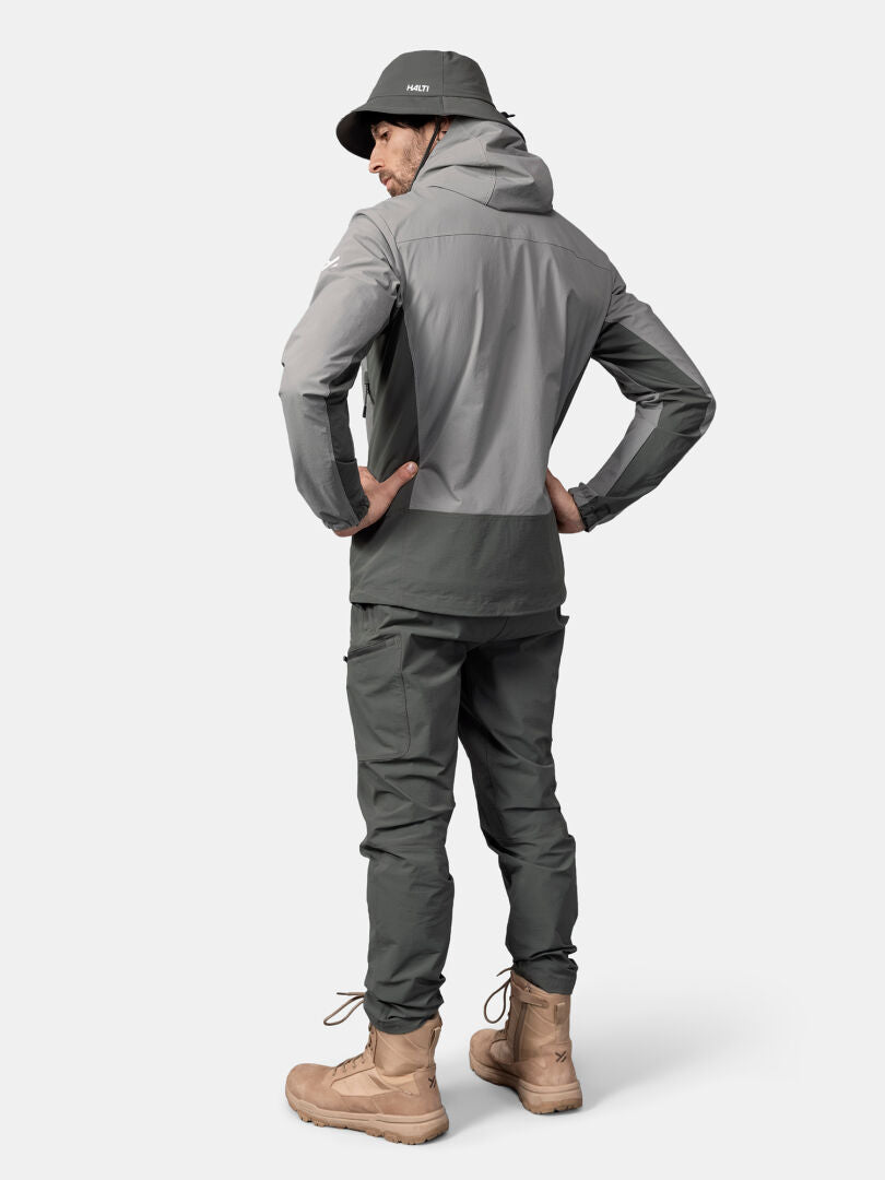 Jemand in einer grauen Halti Pallas Evo X-stretch Jacke Herren, einer Hose und einem Eimerhut steht mit den Händen in den Hüften und schaut weg.