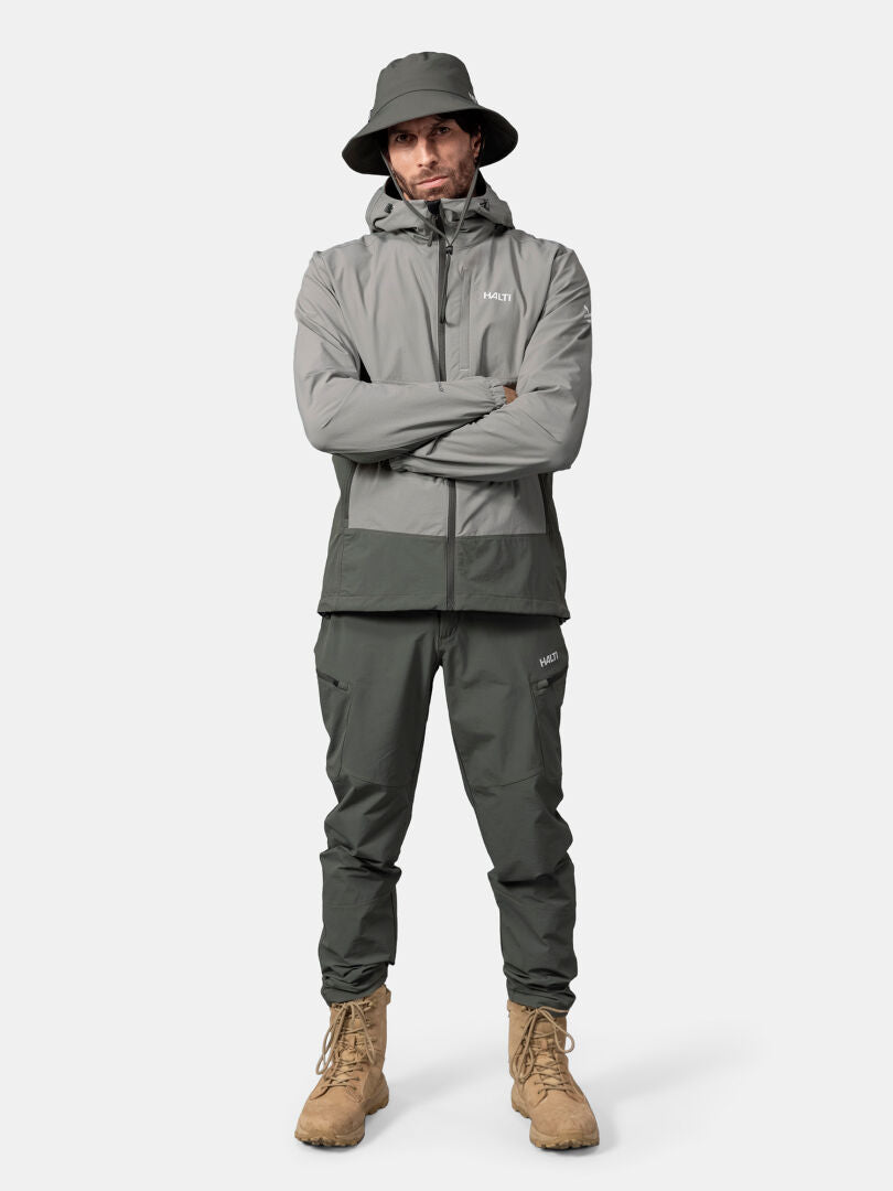 Ein Mann in einer Halti Pallas Evo X-stretch Jacke Herren mit passender Hose, Eimerhut und hellbraunen Stiefeln steht mit verschränkten Armen da.