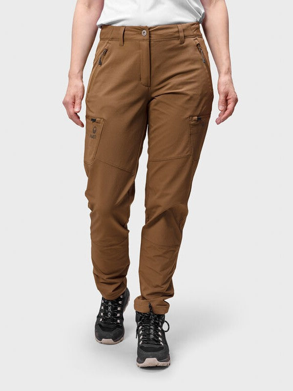 Model trägt Halti Pallas Evo Brushed X-stretch Hosen Damen in braun mit schwarzen Wanderschuhen, auf hellgrauem Hintergrund.