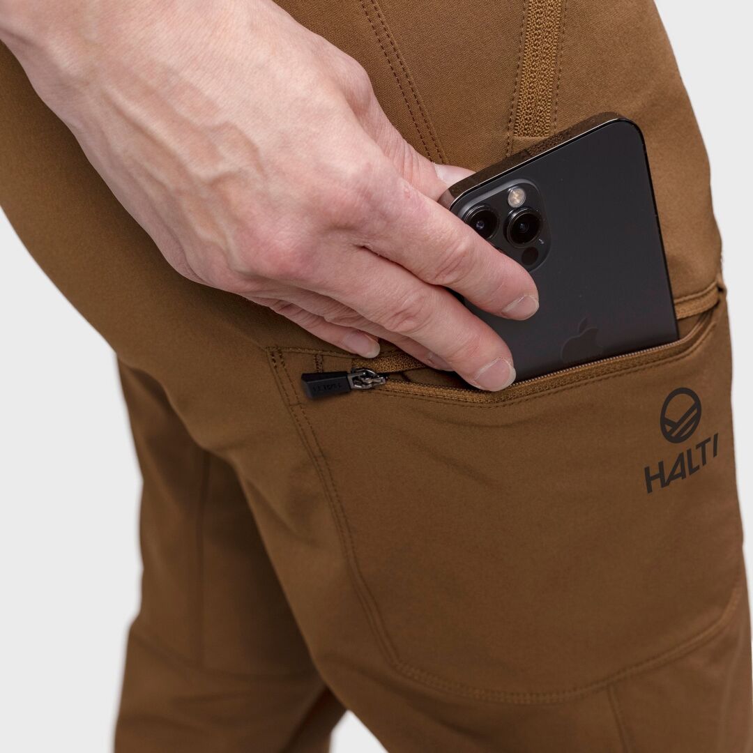 Eine Hand steckt ein schwarzes Smartphone in die Reißverschlusstasche der Halti Pallas Evo Brushed X-Stretch Hosen Damen.