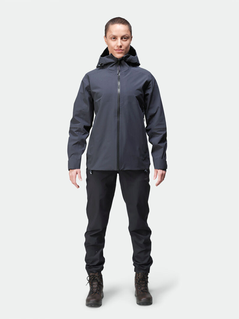 Eine Frau trägt die Halti Kajana 3L DX Regenjacke vor hellem Hintergrund, dazu schwarze Hosen und Stiefel.