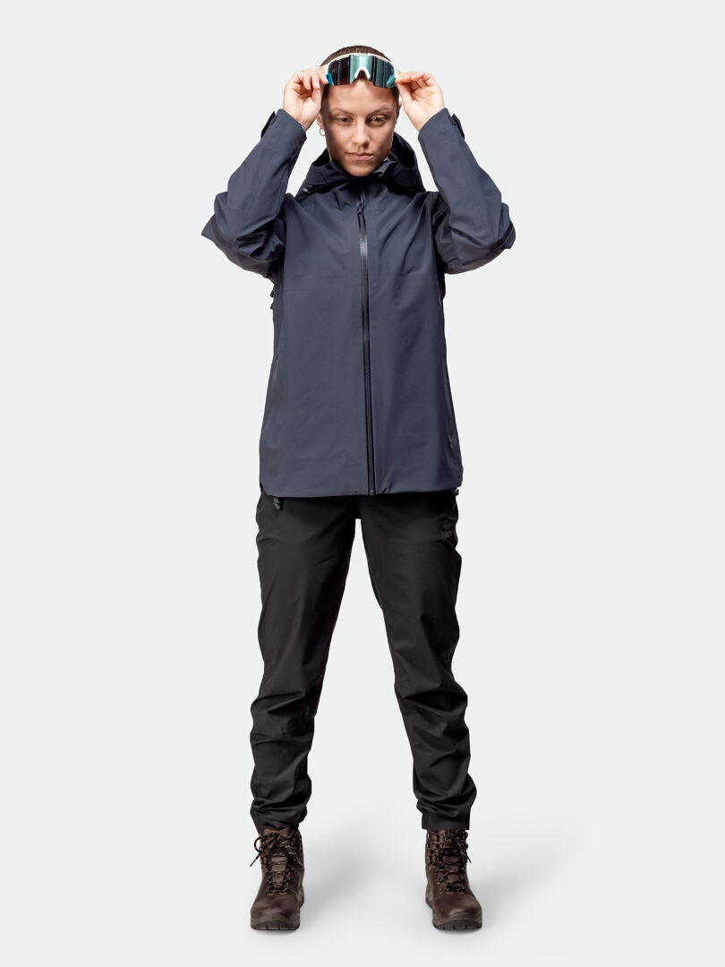 Person trägt die Halti Kajana 3L DX Regenjacke Damen, schwarze Hose, Boots und Ski-Goggles vor neutralem Hintergrund.