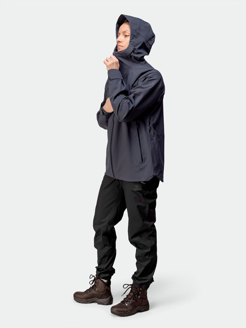 Person trägt die Halti Kajana 3L DX Regenjacke Damen, schwarze Hose und braune Boots vor schlichtem Hintergrund.