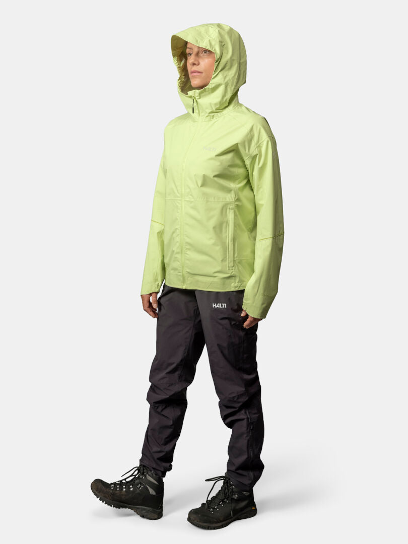 Eine Person in hellgrüner Halti Pisarat 2,5L DX Regenjacke Damen, schwarzer Hose und Wanderschuhen steht vor einem schlichten Hintergrund.