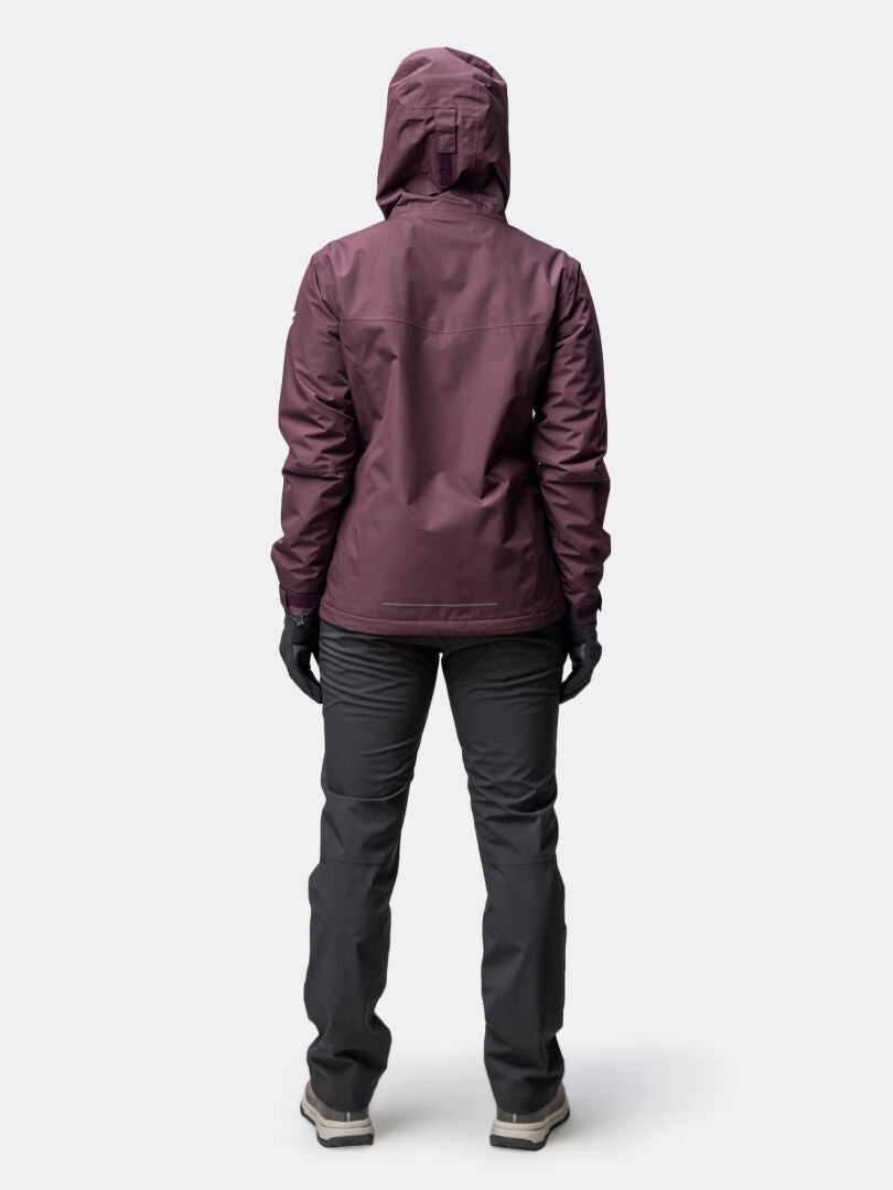 Eine Frau in einer kastanienbraunen Halti Forter DX Warm Regenjacke steht mit dem Gesicht zur Seite auf einem schlichten Hintergrund.