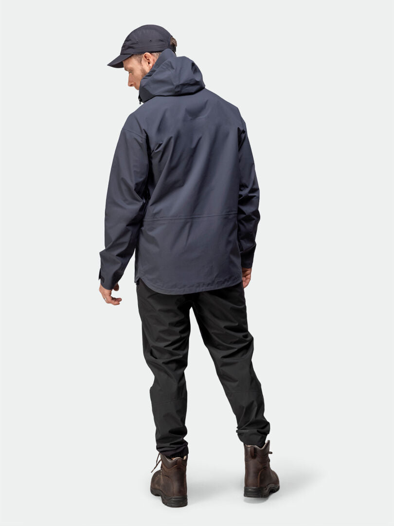 Ein Mann in einer Halti Kajana 3L DX Regenjacke, schwarzen Hosen und braunen Stiefeln blickt abwärts – ideal für Outdoor-Fans.