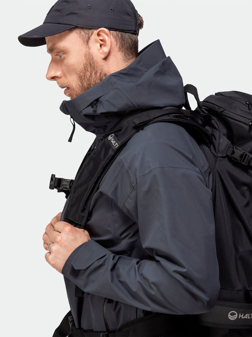 Ein Mann in einer Halti Kajana 3L DX Regenjacke und Mütze trägt einen schwarzen Rucksack und ist bereit für ein Abenteuer im Freien.
