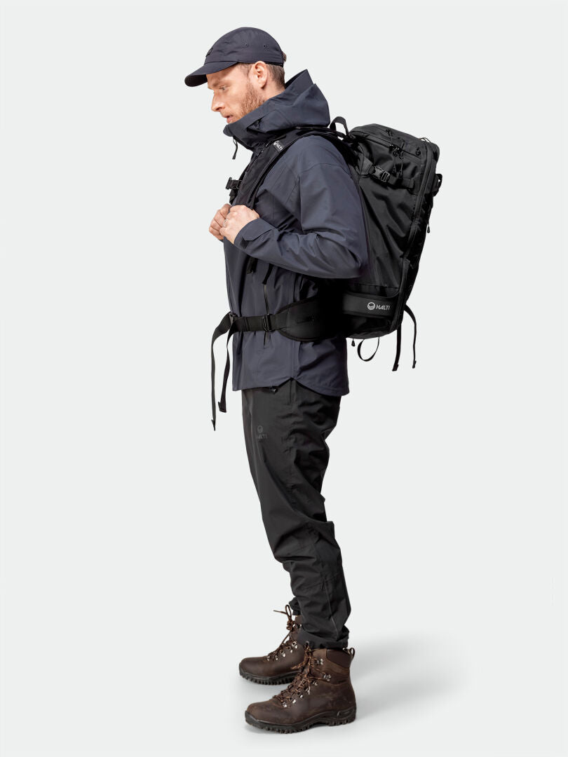 Im Profil steht ein Mann in Outdoor-Ausrüstung, er trägt die Halti Kajana 3L DX Regenjacke für Herren, Hose, Stiefel, Mütze und einen großen Rucksack.