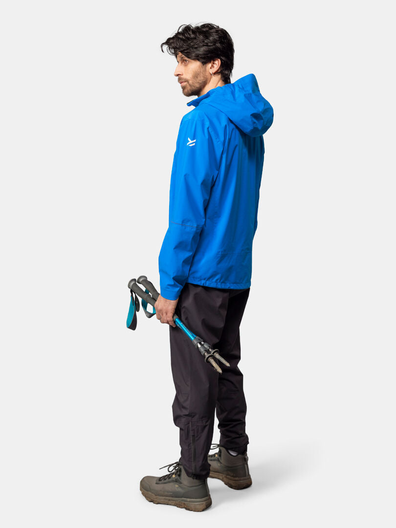 Mann in Halti Pisarat 2,5L DX Regenjacke, schwarzer Hose und Boots mit Trekkingstöcken vor schlichtem Hintergrund.